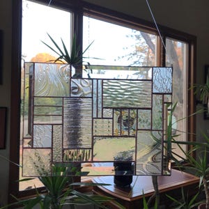 May include: A rectangular stained glass panel with a variety of textured glass pieces in clear, frosted, and patterned designs. The glass is framed with a dark brown border. The panel is hanging in front of a window with plants in the foreground.