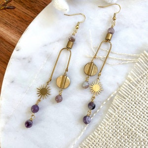 Puede incluir: Un par de pendientes de tono dorado con cuentas de piedras preciosas moradas y grises y abalorios con forma de estrella. Los pendientes cuelgan de una superficie de mármol blanco.