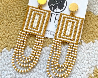 May include: A pair of long, beaded earrings with a geometric design. The earrings are made with white and mustard yellow beads and have gold-toned metal accents.