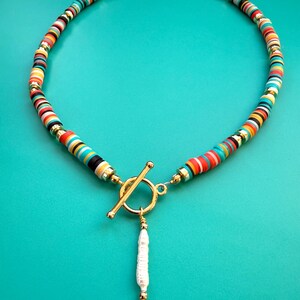 May include: A colorful necklace featuring a strand of flat, round beads in various colors, including red, yellow, blue, and black. The necklace has gold-colored accents and a gold toggle clasp. A white, cylindrical pendant hangs from the clasp.