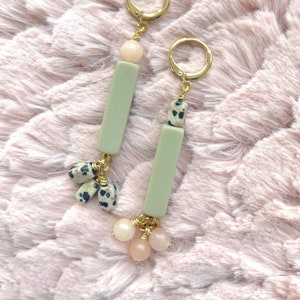 Puede incluir: Un par de pendientes colgantes con cuentas de piedra verde y blanca y cuentas rosas. Los pendientes tienen aros dorados.