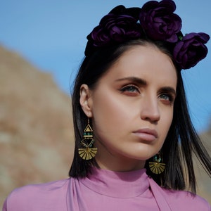 Pode incluir: Uma mulher com cabelo escuro usando uma coroa de flores roxas e brincos de ouro com detalhes verdes. Ela está vestindo uma blusa rosa.