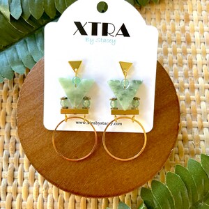 May include: Gold earrings with green gemstone accents and a geometric design. The earrings feature a small hoop and a triangular pendant with a gold bar.