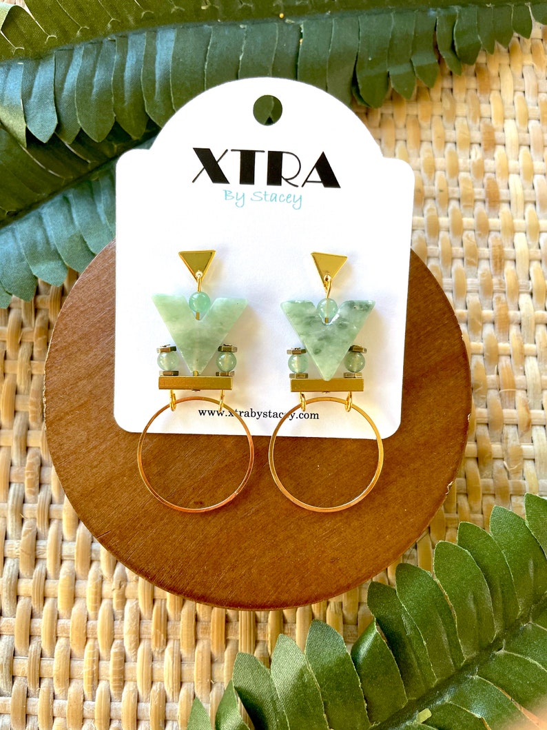 May include: Gold earrings with green gemstone accents and a geometric design. The earrings feature a small hoop and a triangular pendant with a gold bar.