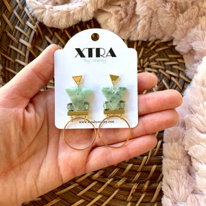 May include: Green and gold geometric earrings with a triangle shape and a hoop design. The earrings are on a white card with the text "XTRA by Stacey" and a website address.