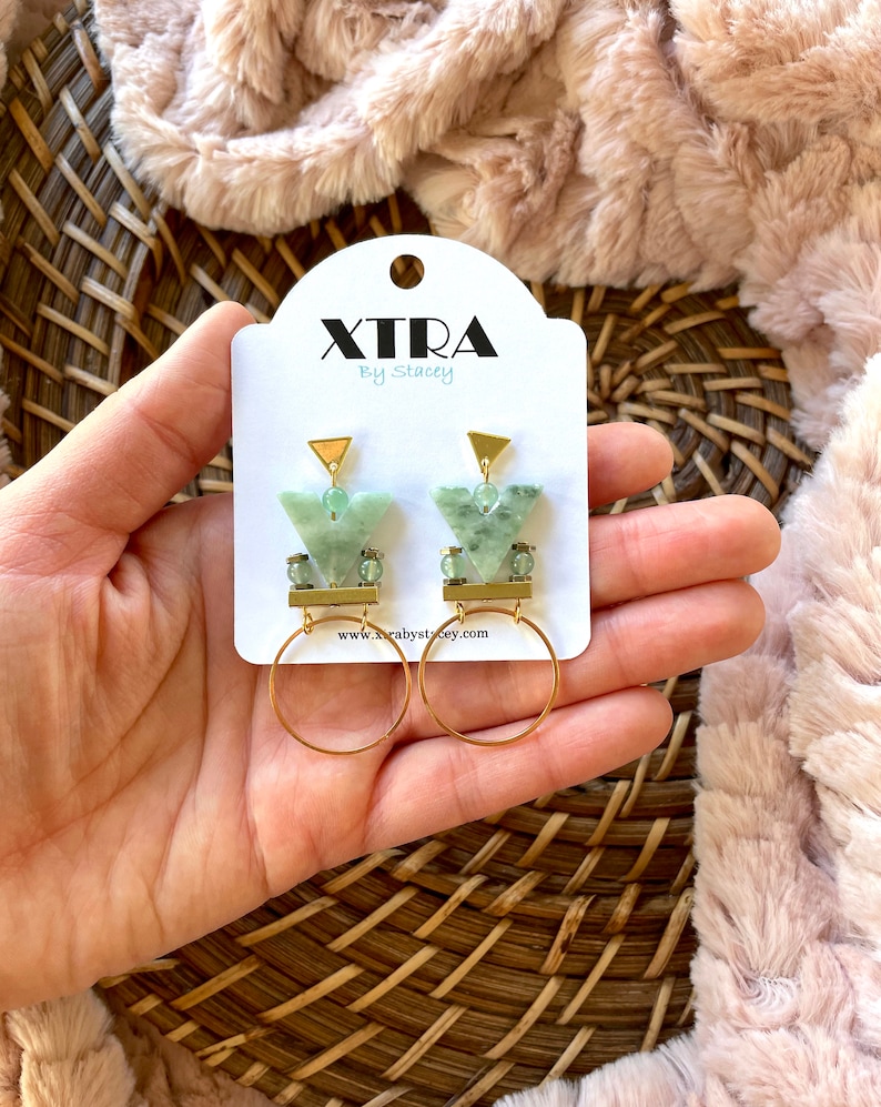 May include: Green and gold geometric earrings with a triangle shape and a hoop design. The earrings are on a white card with the text "XTRA by Stacey" and a website address.