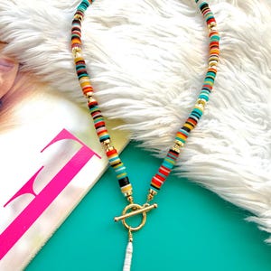 May include: A colorful necklace featuring a toggle clasp and a dangling pearl pendant. The necklace is made of small, flat, round beads in various colors, including red, yellow, blue, and black, separated by gold-colored spacers.