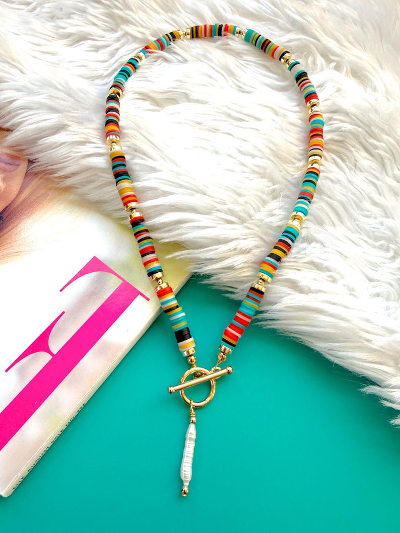 May include: A colorful necklace featuring a toggle clasp and a dangling pearl pendant. The necklace is made of small, flat, round beads in various colors, including red, yellow, blue, and black, separated by gold-colored spacers.