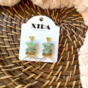 May include: A pair of gold hoop earrings with green stone and gold accents. The earrings are on a white card with the text "Xtra by Stacey" and a website address.