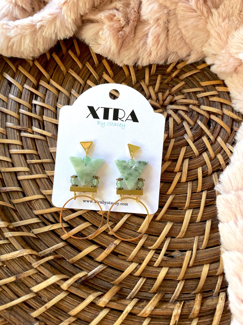 May include: A pair of gold hoop earrings with green stone and gold accents. The earrings are on a white card with the text "Xtra by Stacey" and a website address.