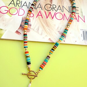 May include: A colorful necklace with a gold clasp and a white pearl pendant. The necklace features a variety of small, flat, round beads in shades of red, orange, yellow, blue, and black. The necklace is displayed on a magazine with the text "ARIANA GRANDE GOD IS A WOMAN."