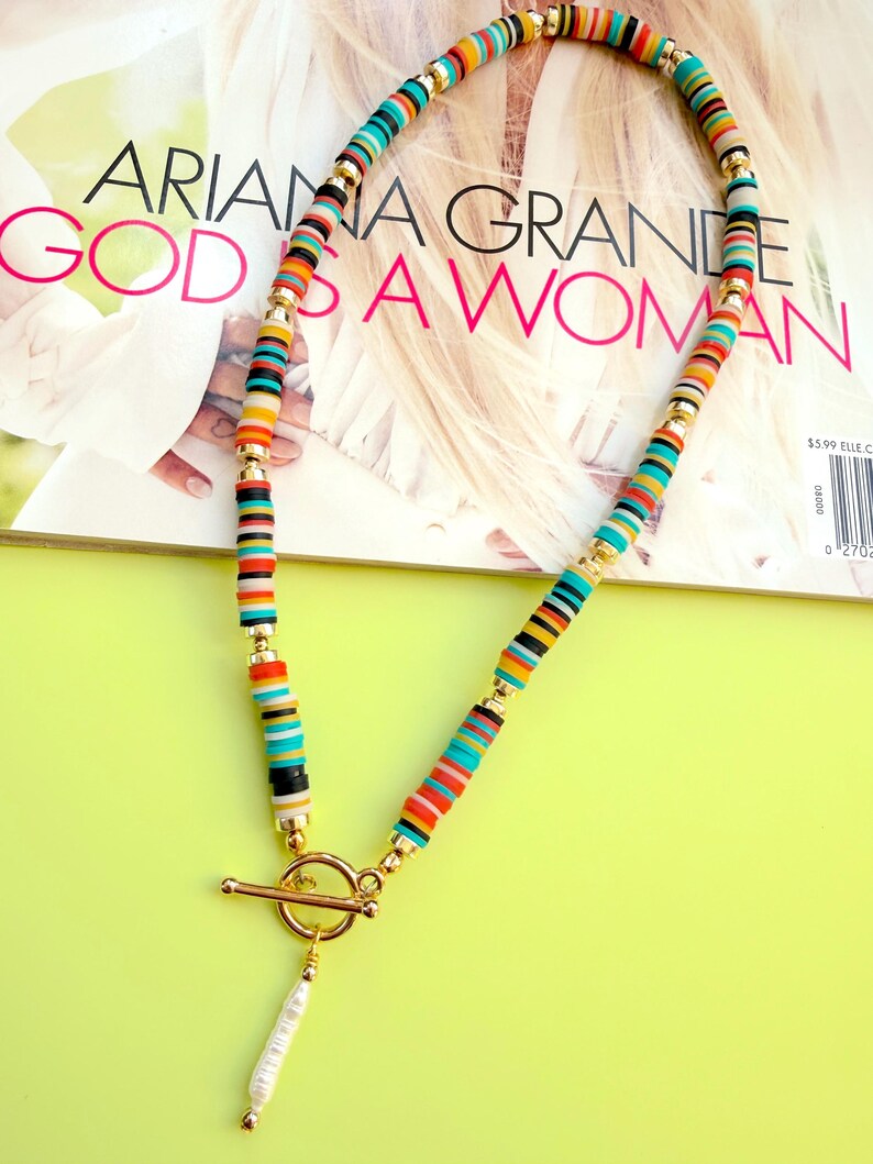 May include: A colorful necklace with a gold clasp and a white pearl pendant. The necklace features a variety of small, flat, round beads in shades of red, orange, yellow, blue, and black. The necklace is displayed on a magazine with the text "ARIANA GRANDE GOD IS A WOMAN."
