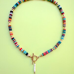 May include: A colorful necklace with a toggle clasp and a dangling pearl pendant. The necklace features a strand of small, flat, round beads in various colors, including blue, red, orange, and black, separated by gold-colored beads.