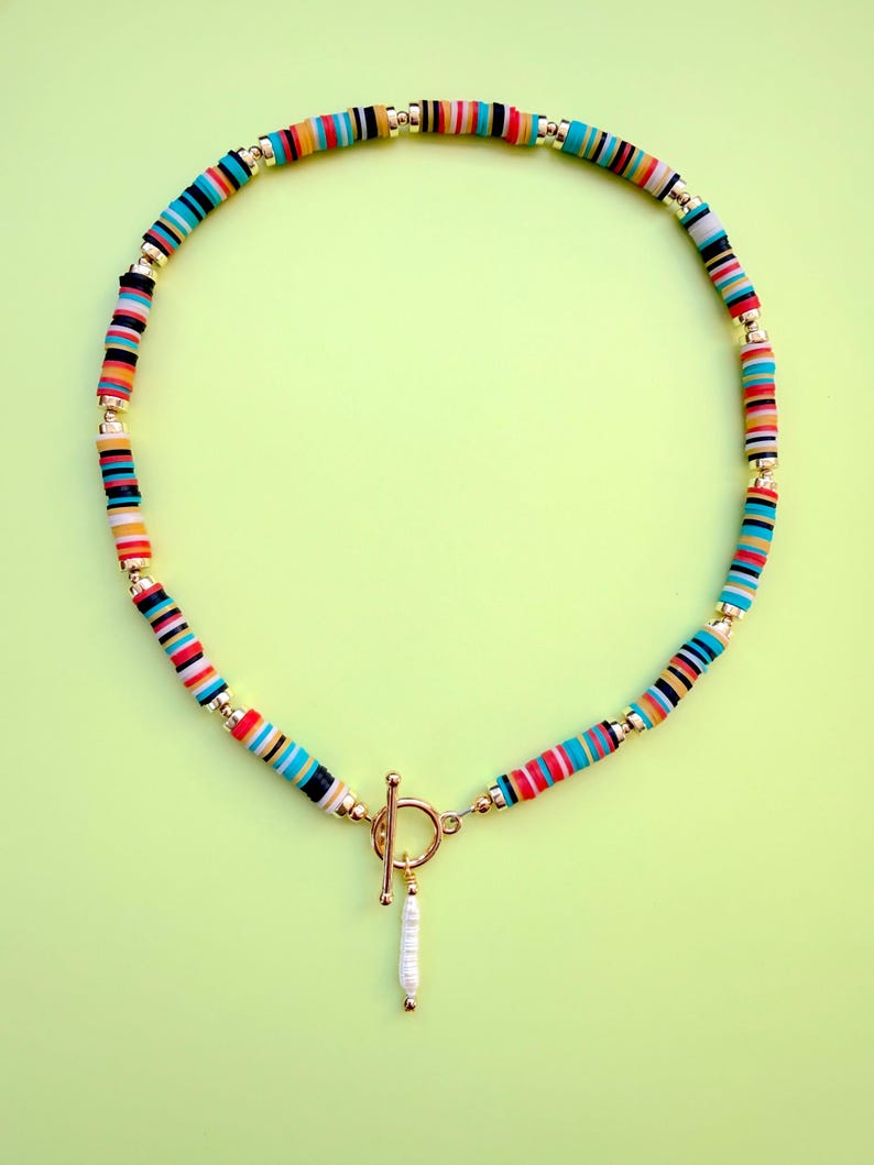 May include: A colorful necklace with a toggle clasp and a dangling pearl pendant. The necklace features a strand of small, flat, round beads in various colors, including blue, red, orange, and black, separated by gold-colored beads.
