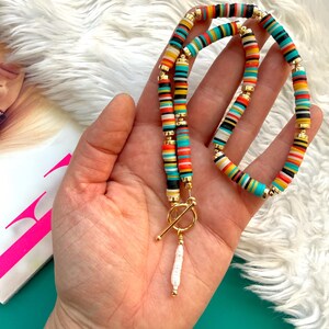 May include: A colorful necklace with a toggle clasp. The necklace features a strand of flat, round beads in various colors, including red, orange, yellow, turquoise, black, and white. A white, elongated pearl pendant hangs from the clasp.