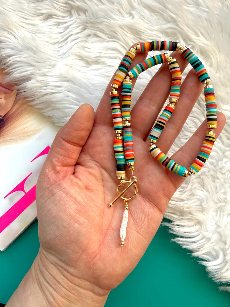 May include: A colorful necklace with a toggle clasp. The necklace features a strand of flat, round beads in various colors, including red, orange, yellow, turquoise, black, and white. A white, elongated pearl pendant hangs from the clasp.