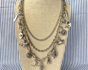 May include: A silver layered chain necklace with multiple charms, including a crescent moon, a skull, a sewing machine, a sun, an octopus, a bird, and a key.