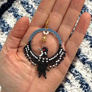 K&ouml;nnte beinhalten: Handgefertigte Perlenohrringe mit einem Vogeldesign. Die Ohrringe haben einen kreisf&ouml;rmigen Reifen mit blauen und wei&szlig;en Perlen, einen goldfarbenen Haken und einen kleinen goldenen Sternanh&auml;nger. Der Vogel ist schwarz und wei&szlig; mit einem roten Auge.