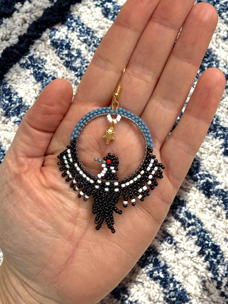 K&ouml;nnte beinhalten: Handgefertigte Perlenohrringe mit einem Vogeldesign. Die Ohrringe haben einen kreisf&ouml;rmigen Reifen mit blauen und wei&szlig;en Perlen, einen goldfarbenen Haken und einen kleinen goldenen Sternanh&auml;nger. Der Vogel ist schwarz und wei&szlig; mit einem roten Auge.