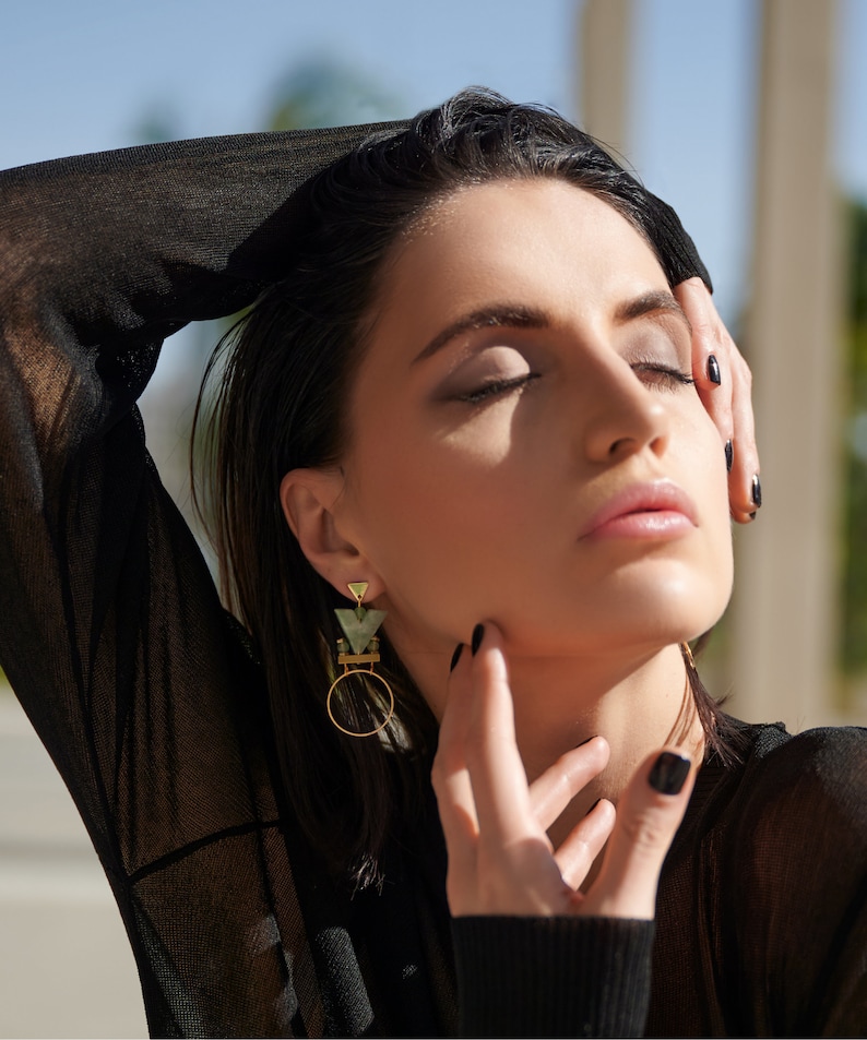 May include: A woman with dark hair wearing a black sheer top and geometric gold and green earrings.
