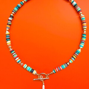 May include: A colorful necklace with a toggle clasp and a white pendant. The necklace features small, flat, round beads in various colors, including red, blue, yellow, black, and white, separated by gold-colored beads. The background is a solid orange.
