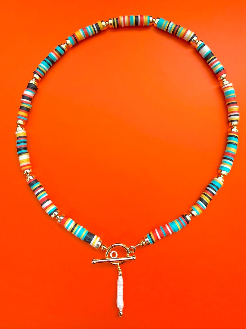 May include: A colorful necklace with a toggle clasp and a white pendant. The necklace features small, flat, round beads in various colors, including red, blue, yellow, black, and white, separated by gold-colored beads. The background is a solid orange.