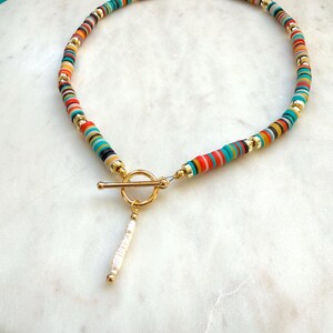 May include: A colorful beaded necklace with a gold-tone toggle clasp. The necklace features small, flat, round beads in various colors, including red, orange, yellow, green, and blue. A white and gold pendant hangs from the clasp.