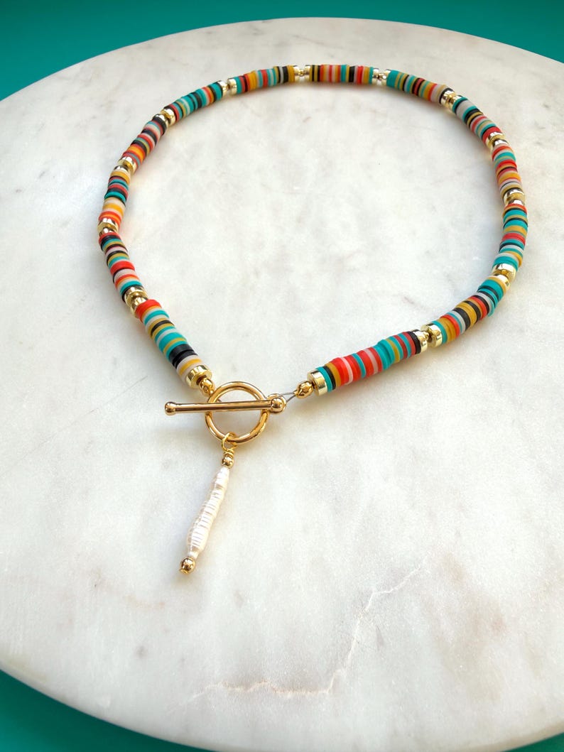 May include: A colorful beaded necklace with a gold-tone toggle clasp. The necklace features small, flat, round beads in various colors, including red, orange, yellow, green, and blue. A white and gold pendant hangs from the clasp.