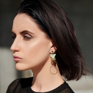 May include: A woman with short dark hair wearing a black shirt and geometric gold and green earrings with a white stone.