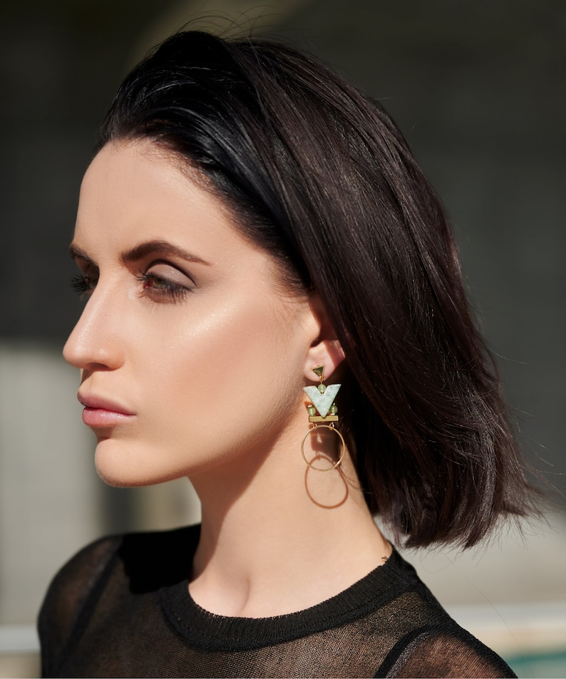 May include: A woman with short dark hair wearing a black shirt and geometric gold and green earrings with a white stone.