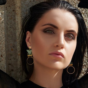 May include: A woman with dark hair and green eyeshadow wears a pair of gold hoop earrings with green geometric accents.