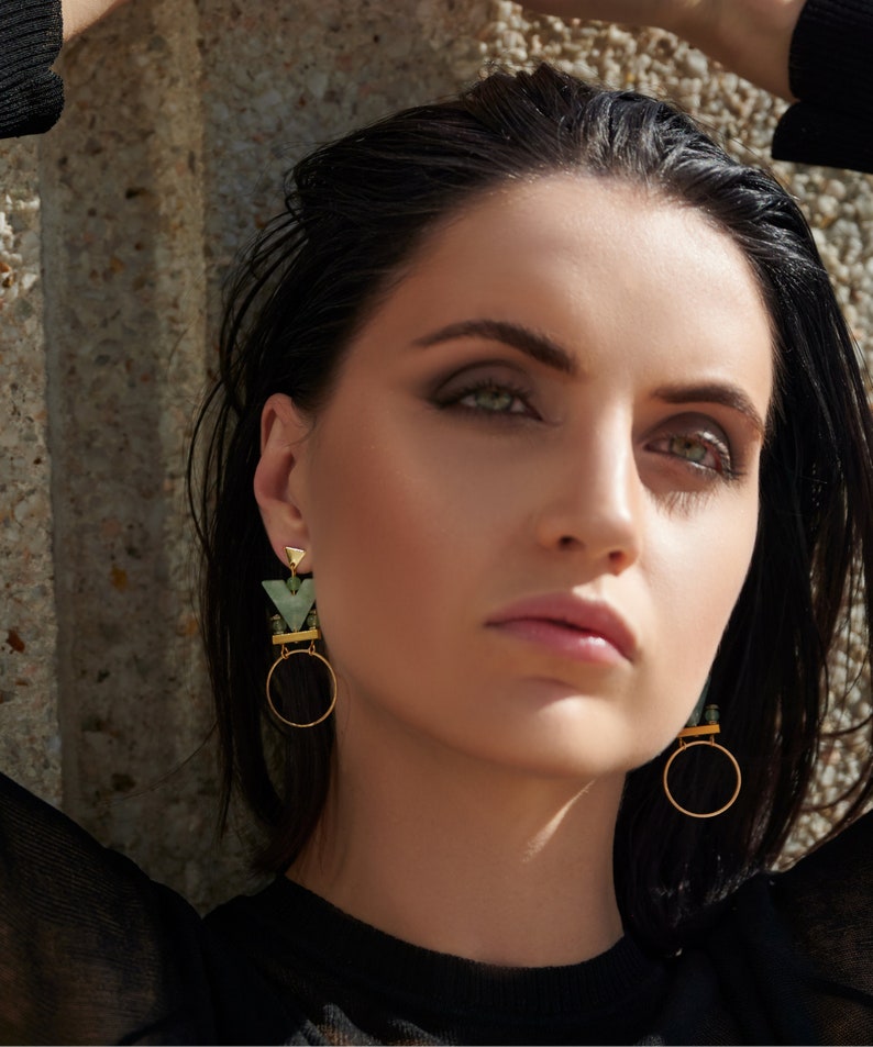 May include: A woman with dark hair and green eyeshadow wears a pair of gold hoop earrings with green geometric accents.