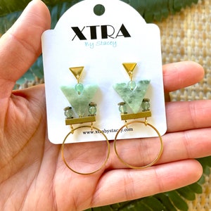 May include: Gold hoop earrings with a green stone pendant. The pendant is a geometric shape with a gold triangle at the top. The earrings are on a white card with the text "XTRA By Stacey" and a website address.