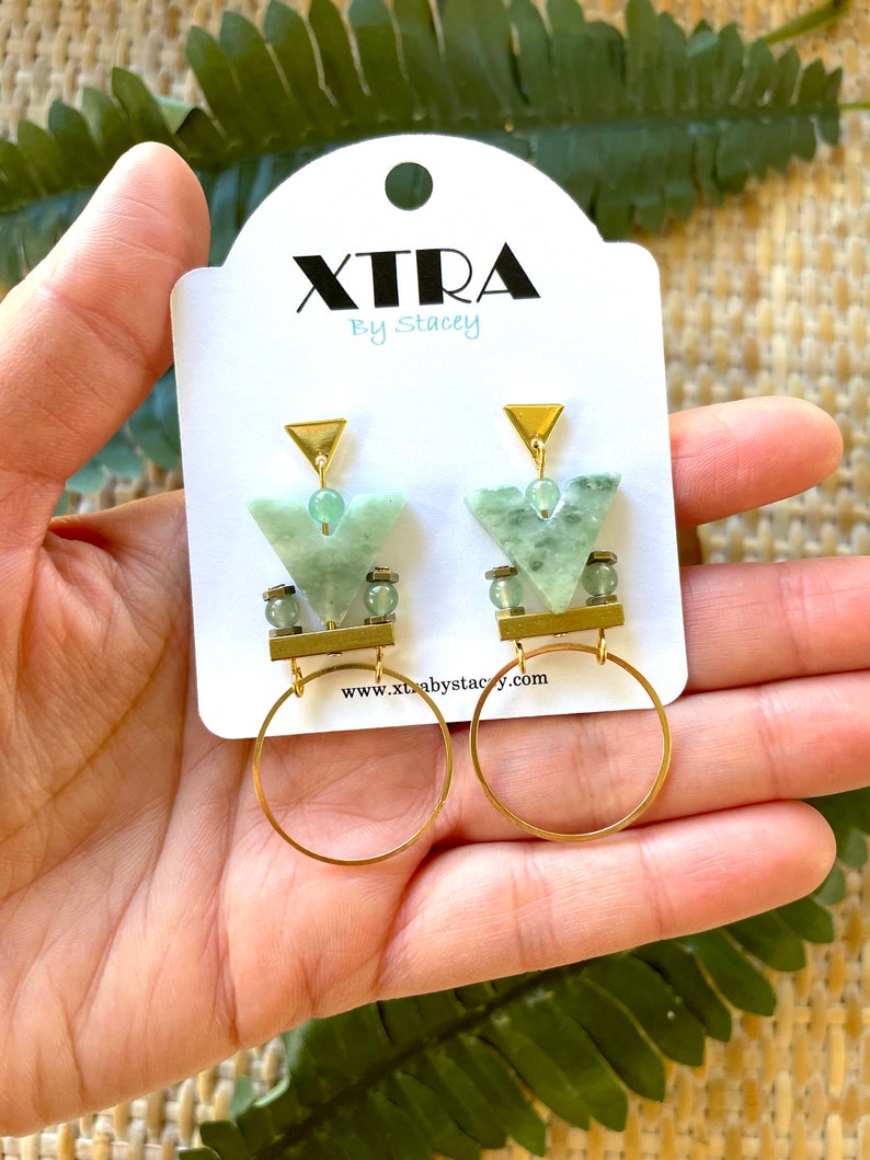 May include: Gold hoop earrings with a green stone pendant. The pendant is a geometric shape with a gold triangle at the top. The earrings are on a white card with the text "XTRA By Stacey" and a website address.