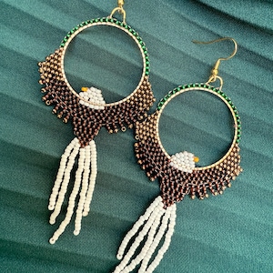 May include: A pair of handcrafted hoop earrings featuring an eagle design. The earrings have gold-colored hoops with green and brown beaded accents. The eagle's body is white and brown, with white beaded fringe.