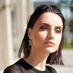 May include: A woman with short dark hair wearing a black shirt and geometric green and gold earrings.