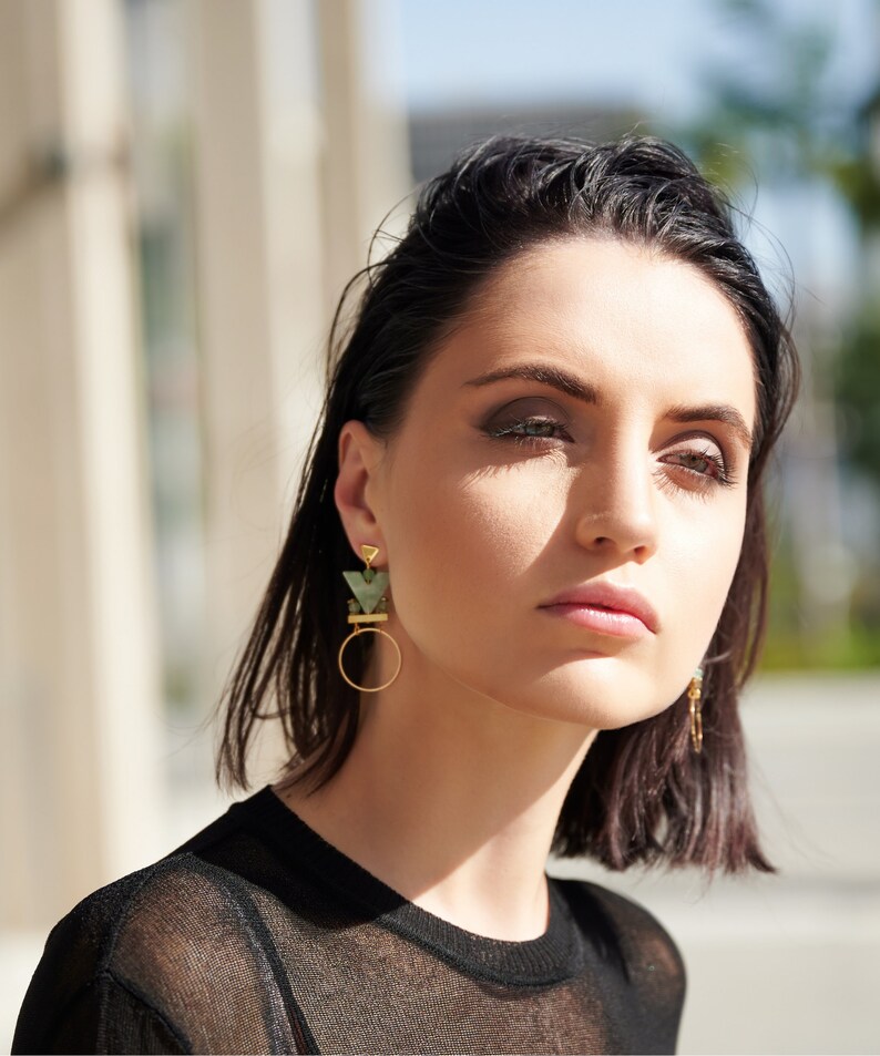 May include: A woman with short dark hair wearing a black shirt and geometric green and gold earrings.
