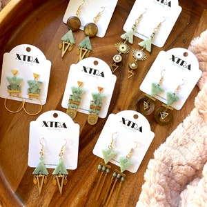 May include: A collection of eight pairs of green and gold geometric earrings on white cards with the brand name "XTRA by stacey".