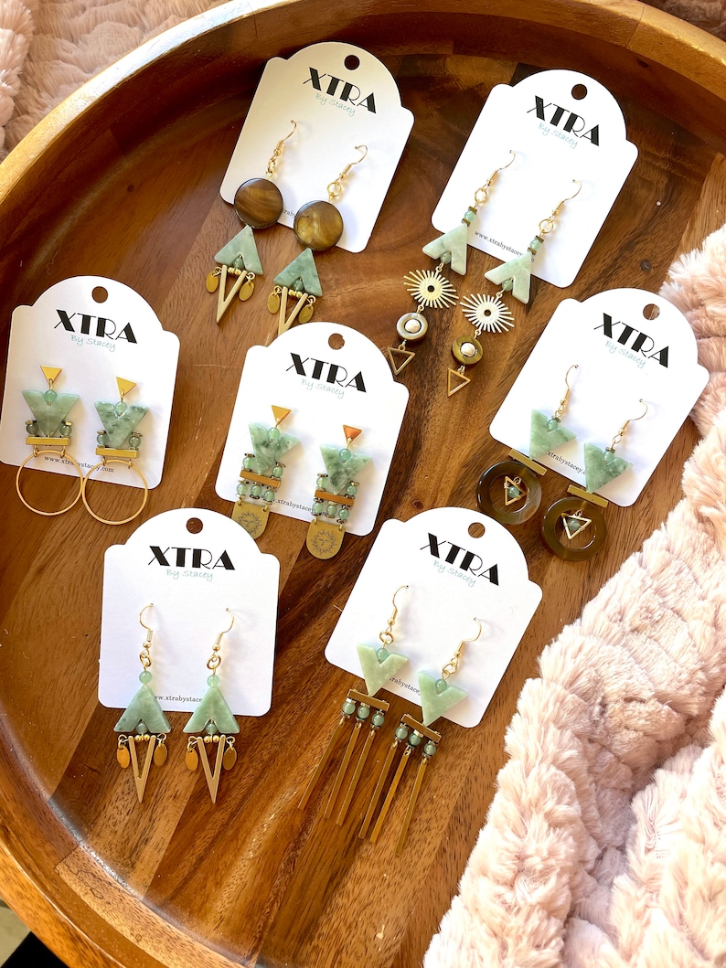 May include: A collection of eight pairs of green and gold geometric earrings on white cards with the brand name "XTRA by stacey".