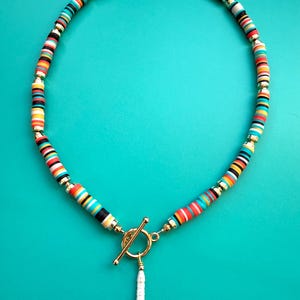 May include: A colorful necklace with a toggle clasp and a white pendant. The necklace features a pattern of small, round, multi-colored beads in shades of red, yellow, blue, black, and white, interspersed with gold-colored beads.