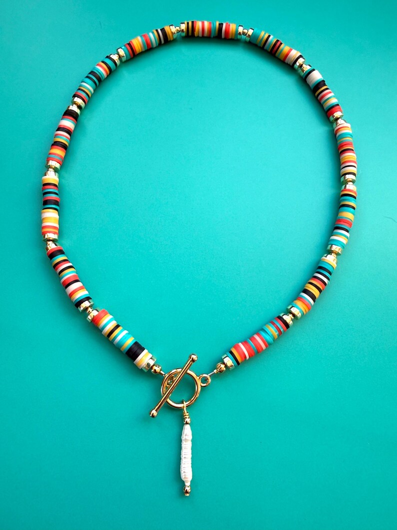 May include: A colorful necklace with a toggle clasp and a white pendant. The necklace features a pattern of small, round, multi-colored beads in shades of red, yellow, blue, black, and white, interspersed with gold-colored beads.