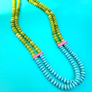 May include: A double-strand necklace featuring round, translucent lime green beads and flat, disc-shaped turquoise blue beads. The two sections are separated by rectangular, solid pink spacers, and the necklace is secured with a gold-tone toggle clasp.