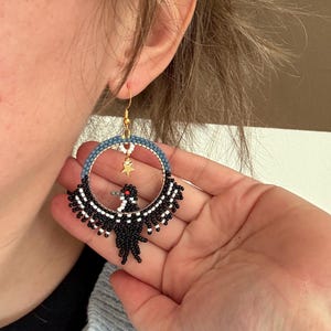 K&ouml;nnte beinhalten: Handgefertigte Creolen-Ohrringe mit einem schwarzen Vogeldesign und wei&szlig;en Akzenten. Der obere Teil des Reifens ist blau und wei&szlig;, mit einem goldenen Sternanh&auml;nger. Die Ohrringe haben goldfarbene Haken.