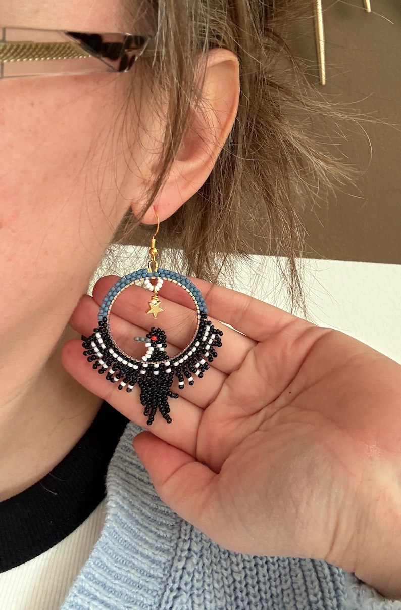 K&ouml;nnte beinhalten: Handgefertigte Creolen-Ohrringe mit einem schwarzen Vogeldesign und wei&szlig;en Akzenten. Der obere Teil des Reifens ist blau und wei&szlig;, mit einem goldenen Sternanh&auml;nger. Die Ohrringe haben goldfarbene Haken.