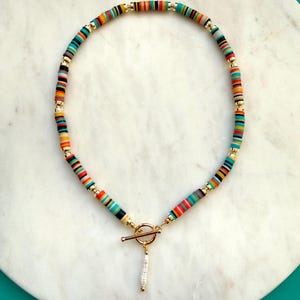 May include: A colorful necklace featuring flat, round beads in a rainbow of colors, including red, orange, yellow, green, and blue. The necklace has gold-colored accents and a toggle clasp with a small, dangling white pendant. The necklace is displayed on a white marble surface.