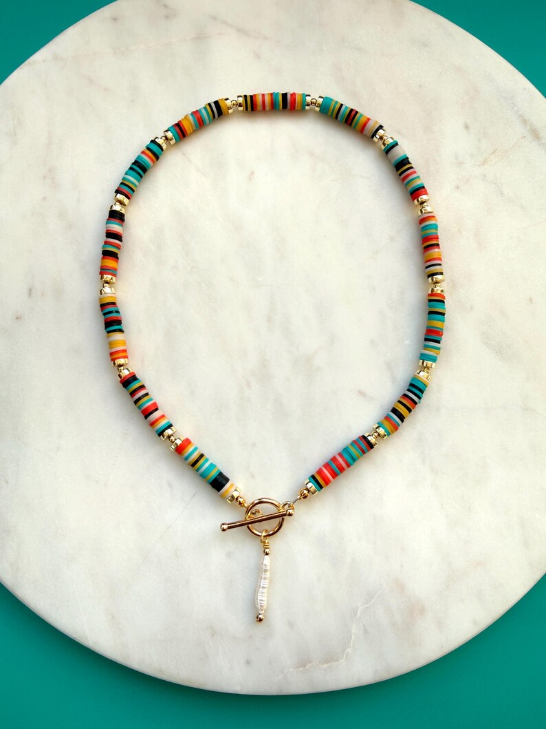 May include: A colorful necklace featuring flat, round beads in a rainbow of colors, including red, orange, yellow, green, and blue. The necklace has gold-colored accents and a toggle clasp with a small, dangling white pendant. The necklace is displayed on a white marble surface.