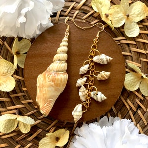 Op de afbeelding: Een paar gouden oorbellen met een schelpdesign. Eén oorbel heeft een grote schelp hanger, terwijl de andere oorbel een ketting met kleinere schelpen heeft.