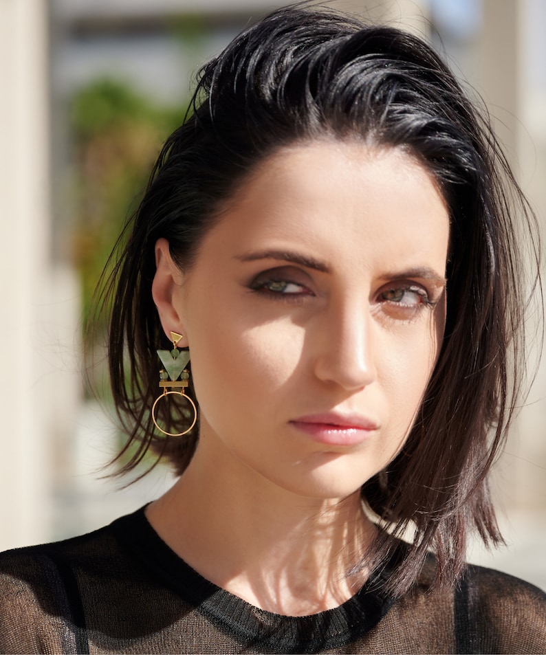 May include: A woman with short black hair wearing a black shirt and geometric green and gold earrings.