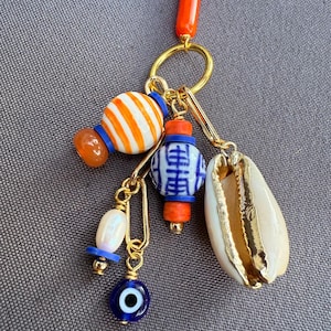 May include: A gold-toned keychain with various charms. Charms include a white and orange striped bead, a blue and white patterned bead, a white seashell with gold trim, a blue evil eye charm, and a white bead. The keychain has an orange handle.