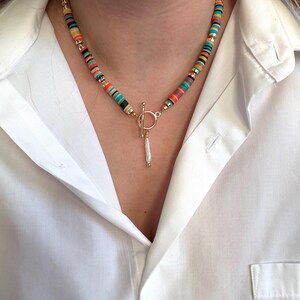 May include: A colorful beaded necklace with a gold clasp and a dangling charm. The necklace features a variety of colors, including red, orange, yellow, green, and blue. The clasp is a gold toggle style. The charm is a small, white, elongated shape.